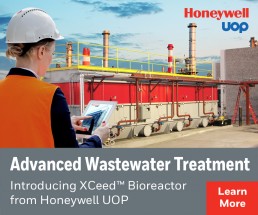 A worker in a hard hat and orange vest uses a tablet near an industrial facility with red machinery. Text: 