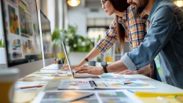 Two professionals collaborate at a desk, focusing intently on computer screens. Charts and graphs are scattered around, conveying a busy, productive atmosphere.